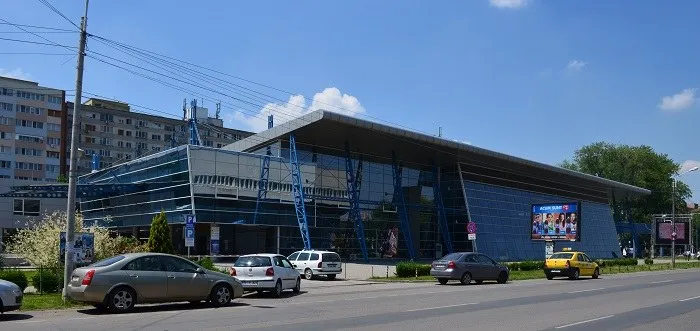 O clădire modernă cu fațadă de sticlă și panouri metalice este situată lângă un drum cu mai multe mașini parcate și în mișcare.