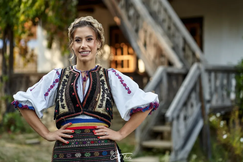 O femeie zâmbindă poartă o cămașă albă cu mâneci largi brodate și o vestă neagră decorată cu motive colorate, stând cu mâinile pe șolduri.