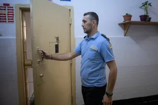 Un bărbat în uniformă de gardian de penitenciar deschide o ușă metalică, în interiorul unei clădiri. Pe perete se află un raft cu plante. 