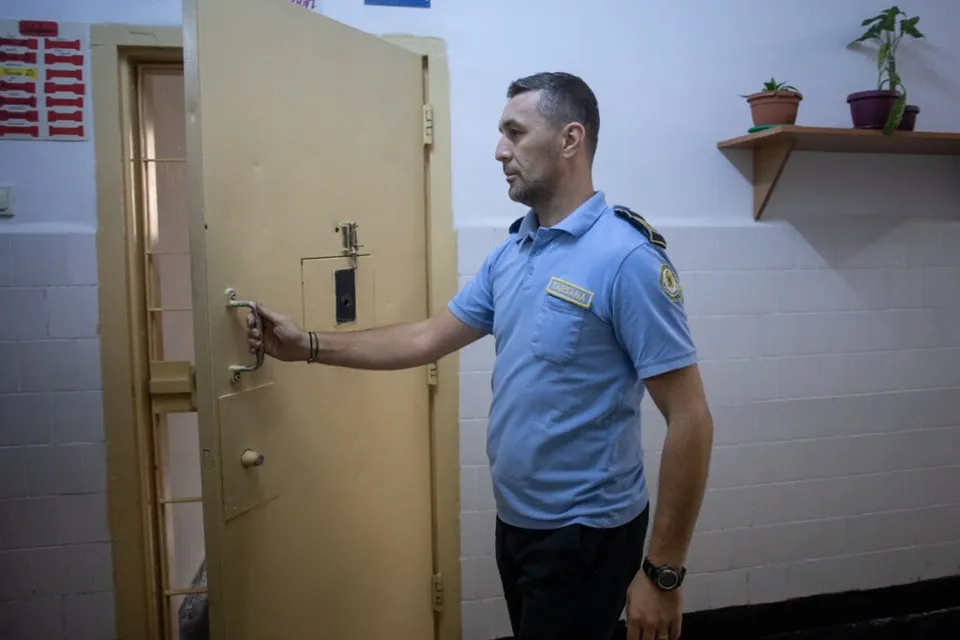 Un bărbat în uniformă de gardian de penitenciar deschide o ușă metalică, în interiorul unei clădiri. Pe perete se află un raft cu plante. 