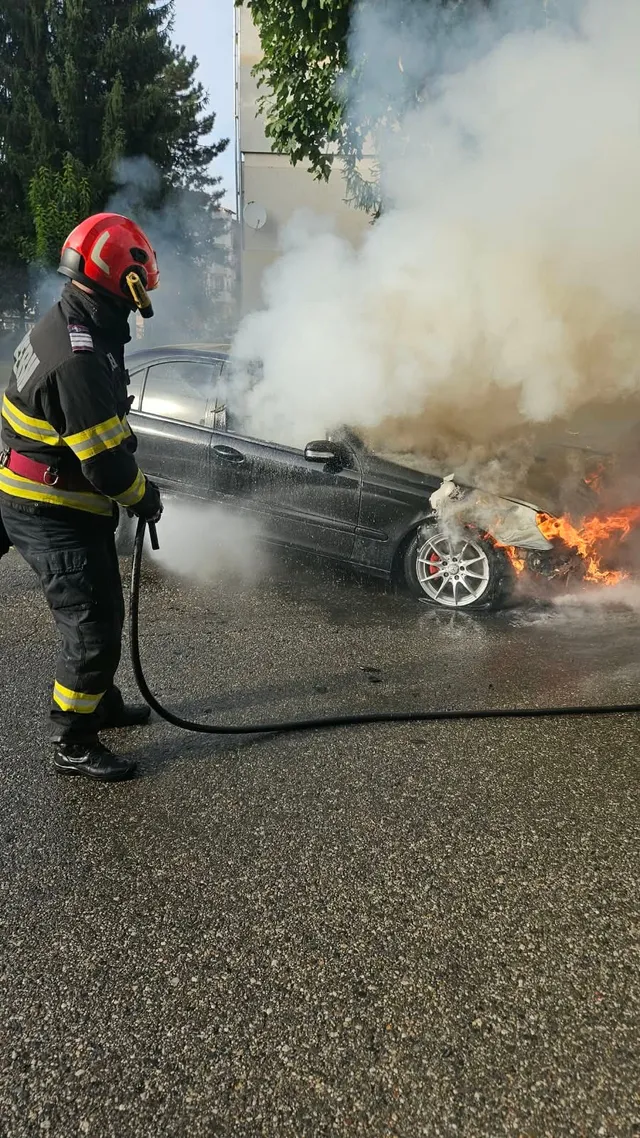 Un pompier cu cască roșie și echipament de protecție folosește un furtun pentru a stinge un autoturism negru din care ies fum și flăcări.