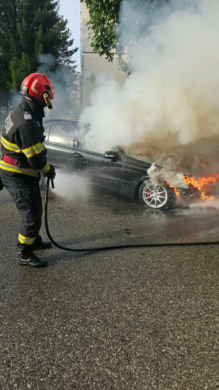 Un pompier cu cască roșie și echipament de protecție folosește un furtun pentru a stinge un autoturism negru din care ies fum și flăcări.