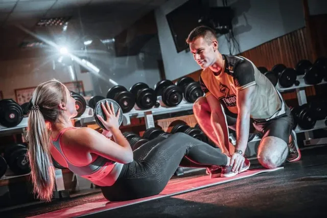 O femeie cu părul blond stă pe un covoraș de yoga ținând o minge de fitness, în timp ce un bărbat stă ghemuit lângă ea într-o sală de sport.