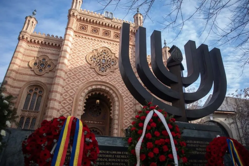 O sculptură metalică în formă de menorah se află în prim-plan, în fața unei clădiri din cărămidă cu elemente arhitecturale decorative și coroane de flori.