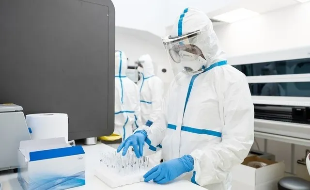 Două persoane purtând costume de protecție albe cu detalii albastre și mănuși de protecție albastre lucrează într-un laborator lângă un monitor negru.