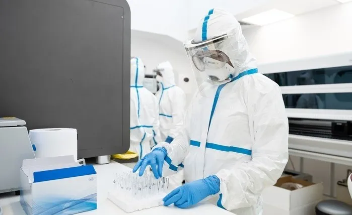 Două persoane purtând costume de protecție albe cu detalii albastre și mănuși de protecție albastre lucrează într-un laborator lângă un monitor negru.