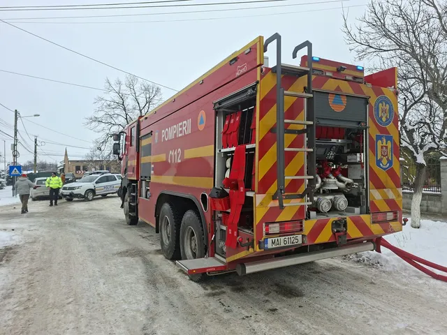 Un autospecială de pompieri roșie cu marcaje galbene este parcată pe un drum de pietriș lângă un autoturism argintiu sub un cer înnorat.