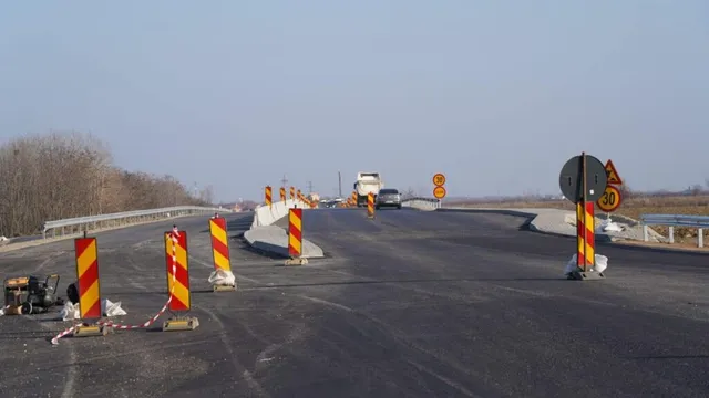 O porțiune de drum asfaltat este delimitată de conuri și bariere portocalii cu dungi galbene, având un vehicul și semne de circulație în fundal.