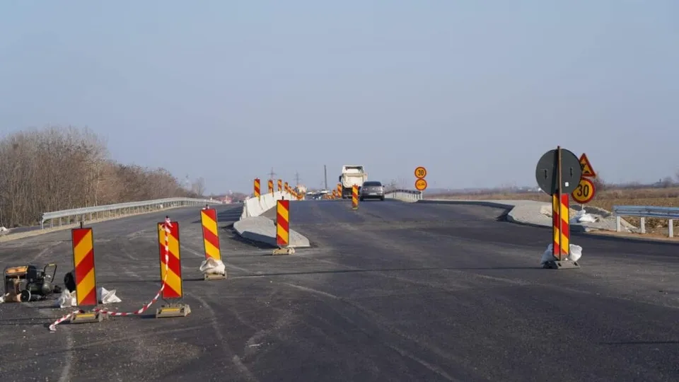 O porțiune de drum asfaltat este delimitată de conuri și bariere portocalii cu dungi galbene, având un vehicul și semne de circulație în fundal.