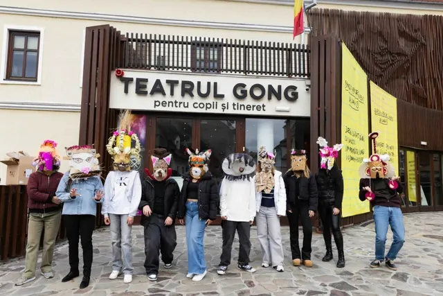 Un grup de adolescenți poartă măști de animale și stă în fața intrării unui teatru cu fațadă albă și elemente din lemn maro.