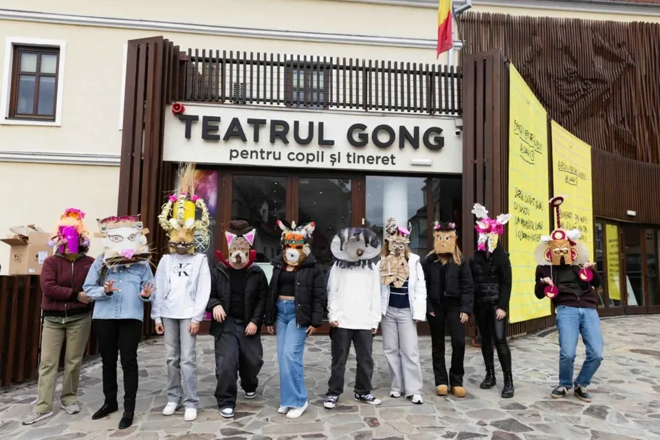 Un grup de adolescenți poartă măști de animale și stă în fața intrării unui teatru cu fațadă albă și elemente din lemn maro.