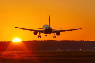 Un avion de pasageri este surprins în zbor, silueta sa fiind vizibilă pe fundalul unui cer portocaliu la apusul soarelui.