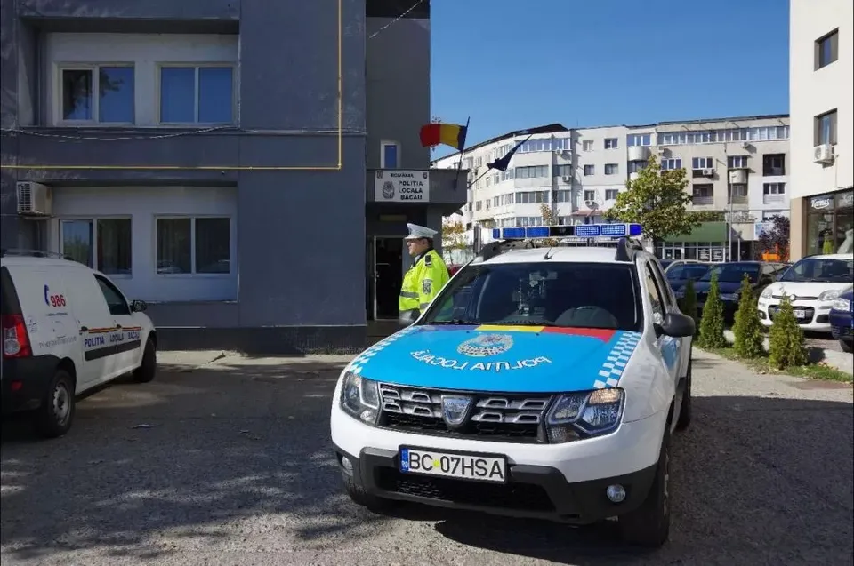 Un vehicul de poliție alb cu detalii albastre și galbene este parcat în fața unei clădiri gri lângă un van alb.