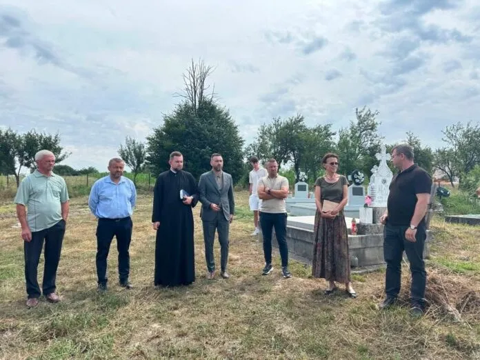 Un grup de opt persoane stă pe un teren cu iarbă lângă un mormânt și câteva monumente funerare sub un cer înnorat.