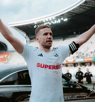 Un bărbat cu mâinile ridicate poartă un tricou alb cu logo-uri și text, stând în fața unor mașini și a unui stadion.