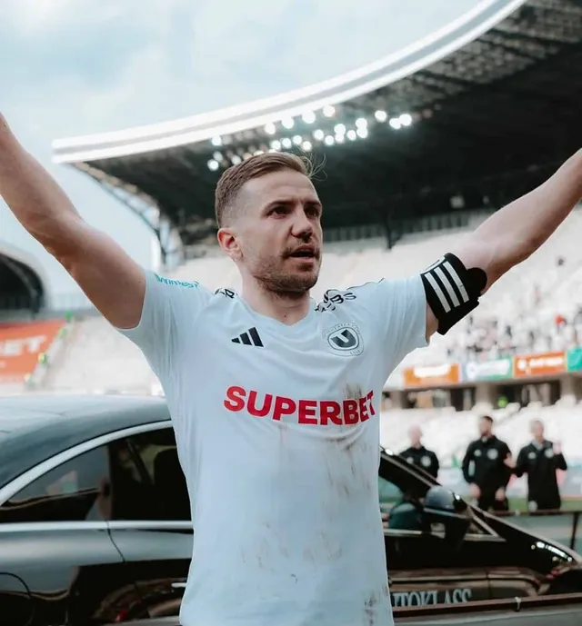 Un bărbat cu mâinile ridicate poartă un tricou alb cu logo-uri și text, stând în fața unor mașini și a unui stadion.