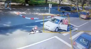O persoană stă întinsă pe asfalt lângă o mașină albă cu portiera deschisă, în timp ce o altă persoană se apleacă spre ea.