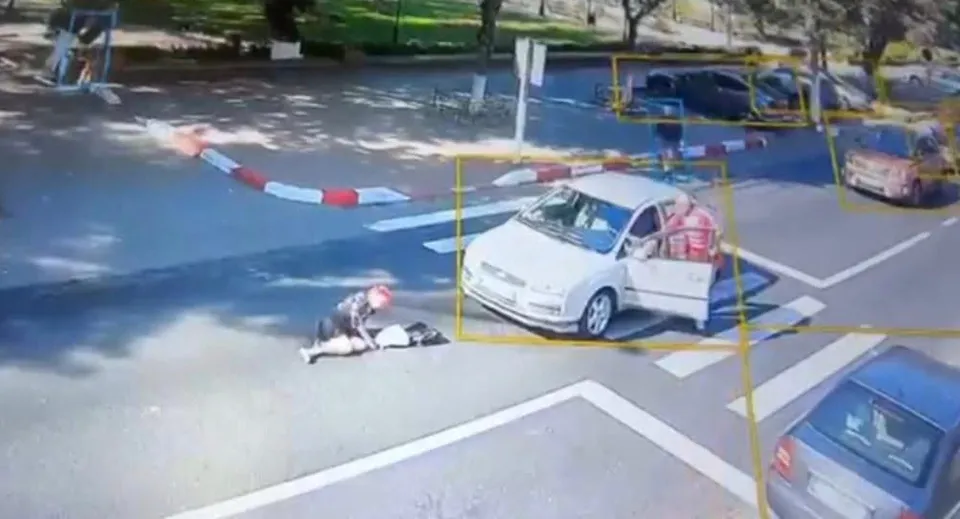 O persoană stă întinsă pe asfalt lângă o mașină albă cu portiera deschisă, în timp ce o altă persoană se apleacă spre ea.