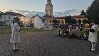 Oameni îmbrăcați în costume istorice albe stau pe un teren pietruit lângă un tun și un tambur, în fața unor clădiri și a unui turn.