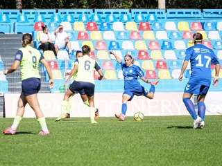 Femei jucând un meci de fotbal pe un teren de iarbă verde, purtând echipamente sportive de culori galben și albastru, în fața unor scaune colorate.