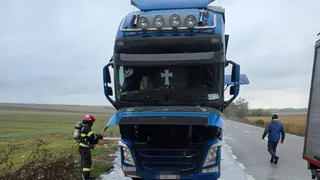 Un camion albastru stă pe marginea unui drum lângă un pompier în echipament de protecție și un om care merge pe asfalt.