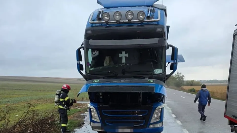 Un camion albastru stă pe marginea unui drum lângă un pompier în echipament de protecție și un om care merge pe asfalt.