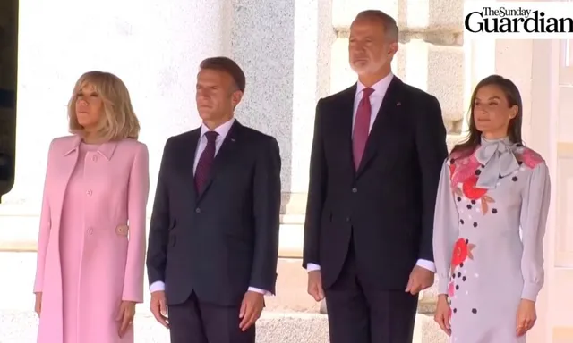 Patru persoane stau în rând, două femei purtând haine roz și gri, iar doi bărbați purtând costume închise și cravate.