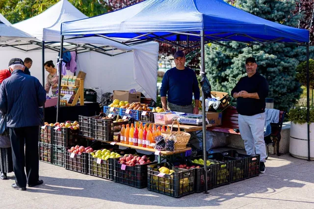 Unii bărbați stau lângă un stand de vânzare acoperit cu un copertină albastră, pe care sunt așezate diverse coșuri cu fructe colorate.