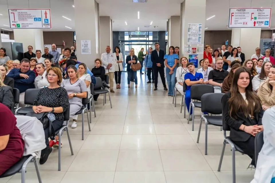 Oamenii stau așezați pe scaune negre într-un hol cu podea deschisă, orientați spre un culoar central unde se află un grup de persoane în picioare.