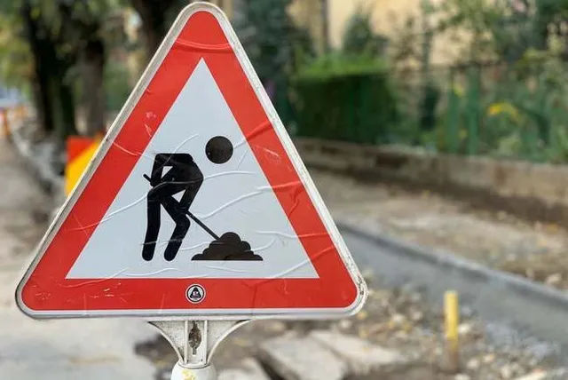Un semn de circulație triunghiular cu margini roșii și un simbol negru de lucrări rutiere este montat pe un stâlp.