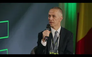 Un bărbat cu părul scurt, îmbrăcat în sacou închis și cămașă albă, ține un microfon negru în mână în fața unui fundal colorat.