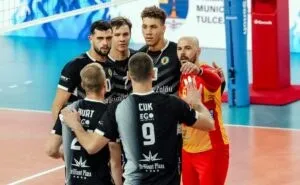 Șase jucători de volei în echipamente negre și unul în echipament roșu și galben stau grupați pe un teren de sport.