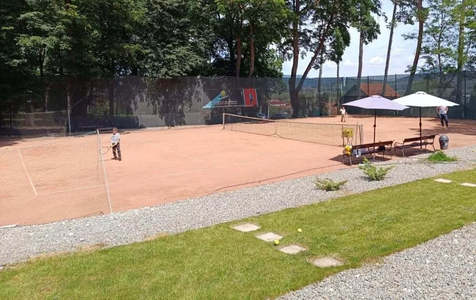Un teren de tenis cu suprafață de zgură, un net întins, două umbrele de grădină și câteva bănci pe marginea terenului.