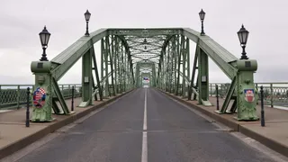 O structură metalică de culoare verde cu parapete și stâlpi de iluminat se întinde peste o cale asfaltată cu o linie albă centrală.