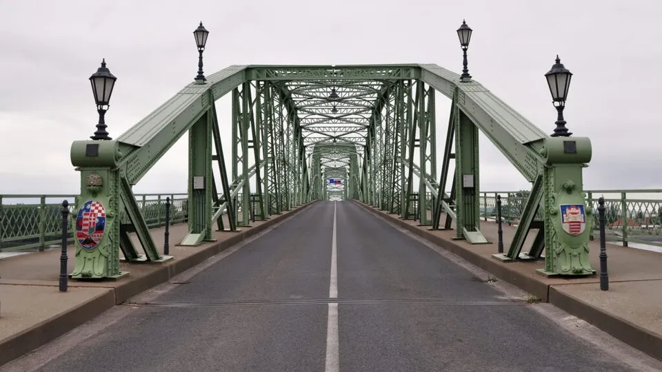 O structură metalică de culoare verde cu parapete și stâlpi de iluminat se întinde peste o cale asfaltată cu o linie albă centrală.