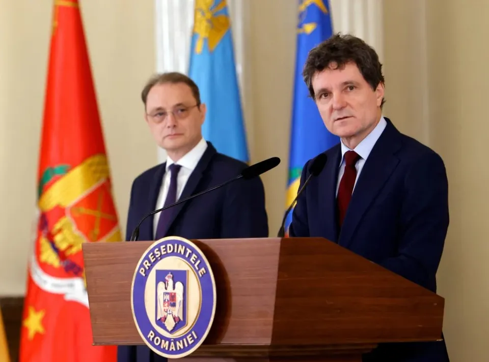 Un bărbat cu ochelari și unul cu cravată roșie stau în spatele unui podium de lemn cu un emblema circulară în fața unor steaguri.