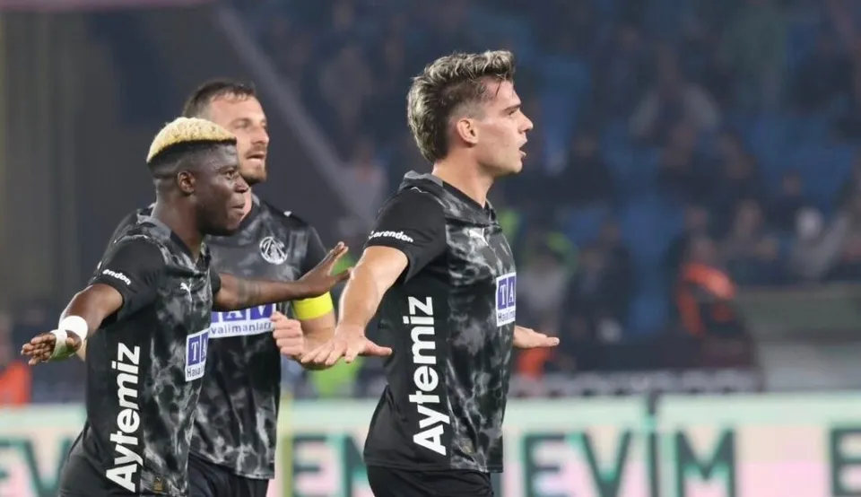 Trei jucători de fotbal în echipamente negre cu inscripții albe sărbătoresc pe un teren de sport sub luminile unui stadion.