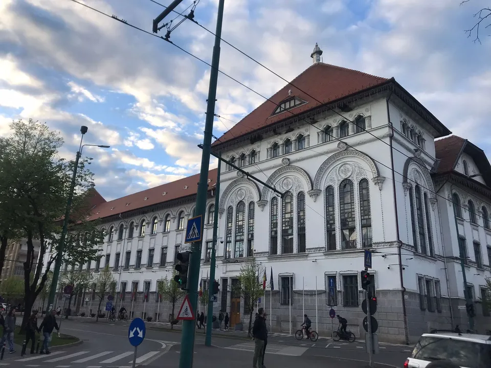 O clădire albă cu acoperiș roșu, ferestre înalte și arcade, situată lângă o stradă cu semne de circulație și câțiva pietoni.