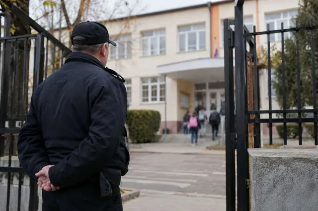 Un bărbat cu șapcă și jachetă neagră stă cu spatele, privind spre intrarea unei clădiri cu ferestre albe și un gard metalic negru.