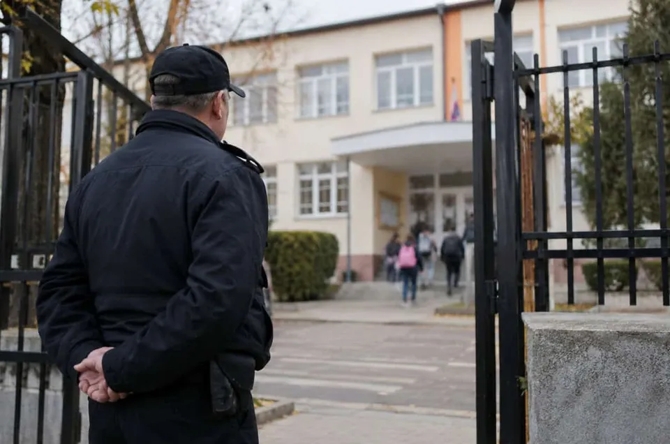 Un bărbat cu șapcă și jachetă neagră stă cu spatele, privind spre intrarea unei clădiri cu ferestre albe și un gard metalic negru.
