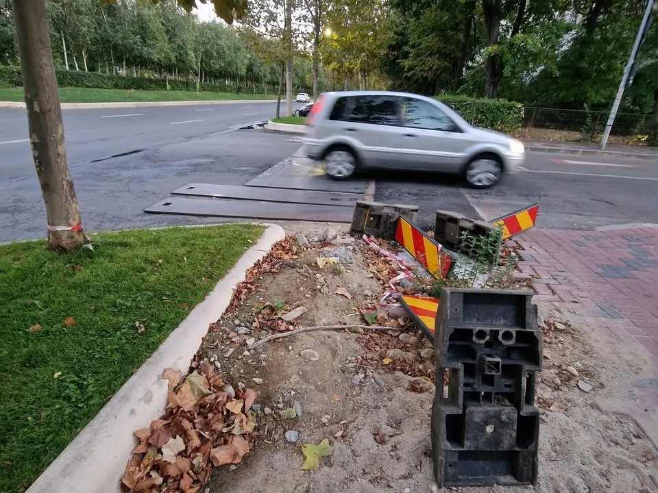 O mașină gri trece peste plăci metalice întinse pe o porțiune de drum lângă un trotuar cu bariere de plastic.
