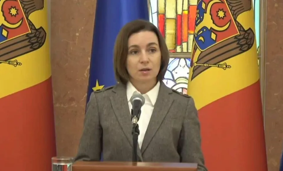 O femeie cu părul scurt și brunet stă în spatele unui podium cu microfon, purtând o jachetă gri și o cămașă albă, în fața unor steaguri.