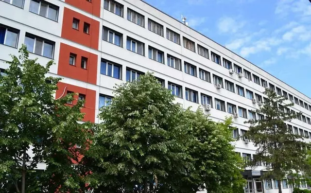 O clădire albă cu ferestre și secțiuni portocalii este parțial acoperită de copaci verzi sub un cer albastru.
