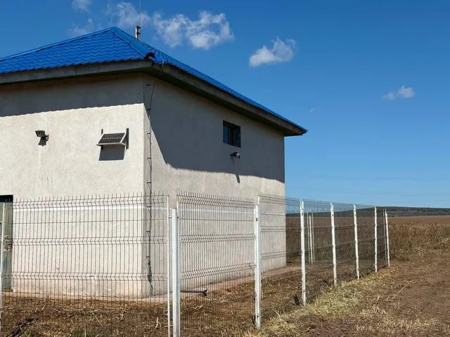 O clădire cu pereți deschiși și acoperiș albastru este înconjurată de un gard metalic pe un teren cu iarbă uscată sub un cer senin.