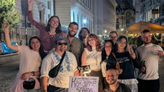 Un grup de oameni zâmbește și ridică mâinile în fața unui afiș care menționează un eveniment de karaoke stradal, într-un decor urban nocturn.