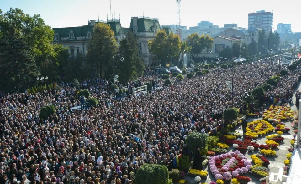 O mulțime mare de oameni este adunată într-o piață publică lângă clădiri și copaci, lângă straturi de flori colorate.