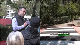 Un bărbat cu vestă gri și cămașă albastră ține un microfon, în timp ce o trecere de pietoni este vizibilă pe asfaltul de lângă vegetație.