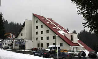 O clădire albă cu acoperiș înclinat de culoare roșie este înconjurată de copaci sub un cer gri, având câteva mașini parcate în fața sa.