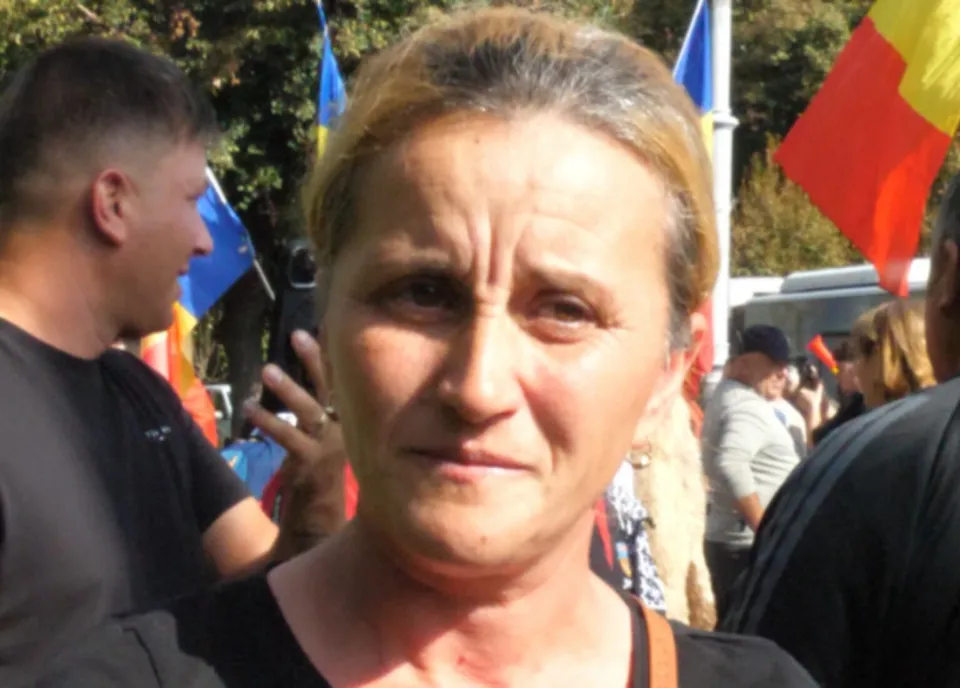 O femeie cu părul strâns în spate privește spre cameră, fiind înconjurată de persoane și steaguri colorate în fundal.