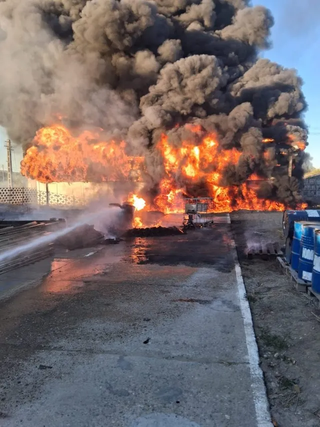 Flăcări mari și un nor dens de fum negru se ridică deasupra unei structuri de depozitare, în timp ce un jet de apă este direcționat spre incendiu.